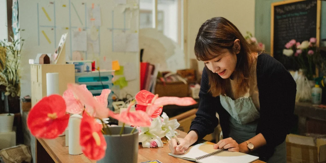 Pessoa anotando um pedido de encomenda em um caderno, com flores ao lado, em ambiente de loja