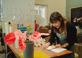 Pessoa anotando um pedido de encomenda em um caderno, com flores ao lado, em ambiente de loja